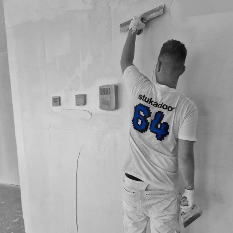 Stukadoor van Stukadoor64 met een spaan aan het werk, met een groot blauw pixel-64 op zijn shirt